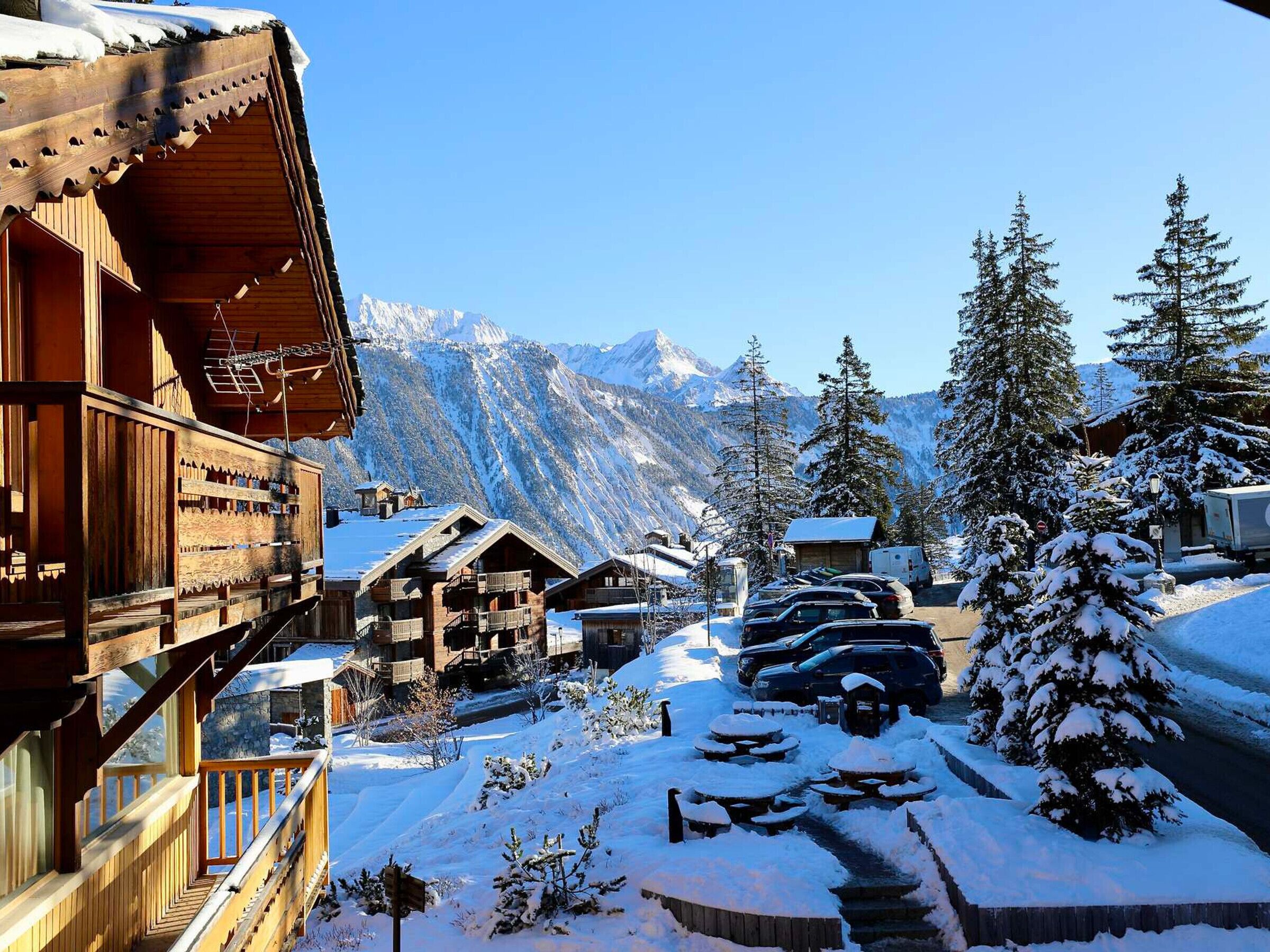 A wooden chalet with balconies, a snow-covered driveway, and a mountain backdrop.