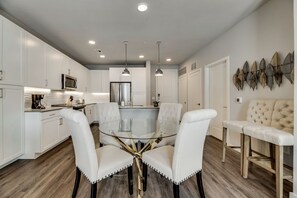 A modern kitchen with white cabinetry, a glass dining table, and pendant lighting. - MCK203 - Apt|Patio|15mi to Toyota Stadium|Sleeps 4 (McKinney)