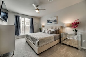 A bedroom with a bed, a ceiling fan, a bedside table with a lamp, a dresser, and a window with curtains.