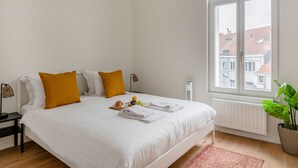 Een netjes opgemaakt bed met witte lakens en twee mosterdgele kussens, een nachtkastje met een lamp, een raam met uitzicht op gebouwen, en een pot met een plant.