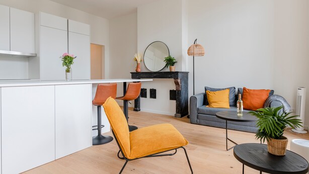 Een moderne keuken met witte kasten, een zwart aanrecht en een gele stoel. Er staat een grijze bank met oranje en blauwe kussens, een klein rond tafeltje en een vloerlamp.