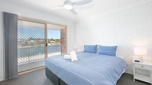 A hotel room with a bed, a nightstand, a lamp, and a view of a body of water through a glass door. - Moondance 2 - Canal front duplex (Yamba)