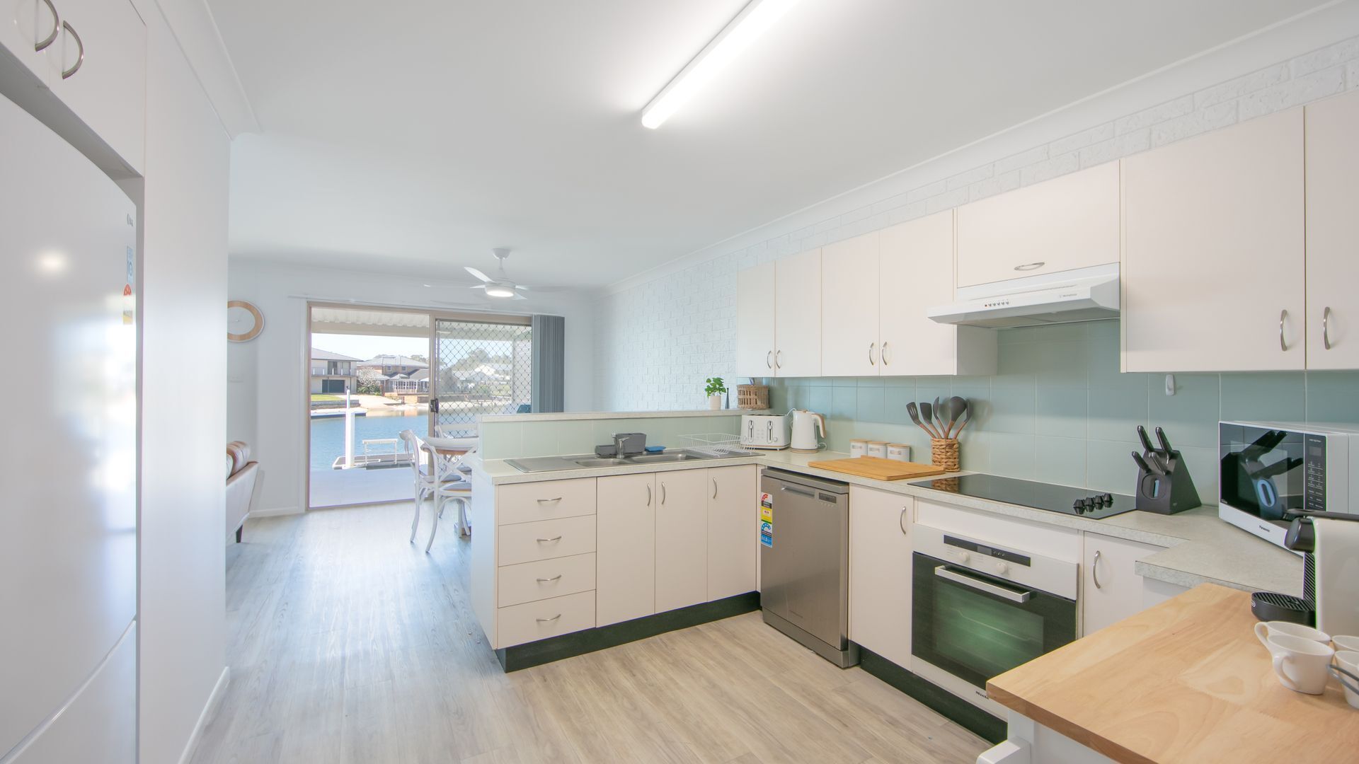 A modern kitchen with white cabinets, a dishwasher, an oven, and a microwave. There is a dining area with a table and chairs visible through a glass door.
