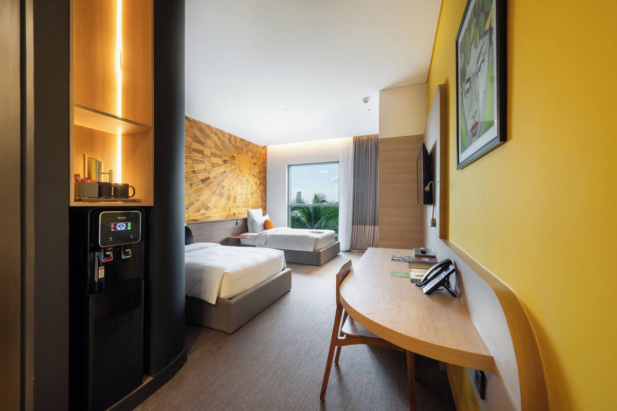 A hotel room with a bed, a desk, a chair, a water dispenser, and a window with a view of greenery.