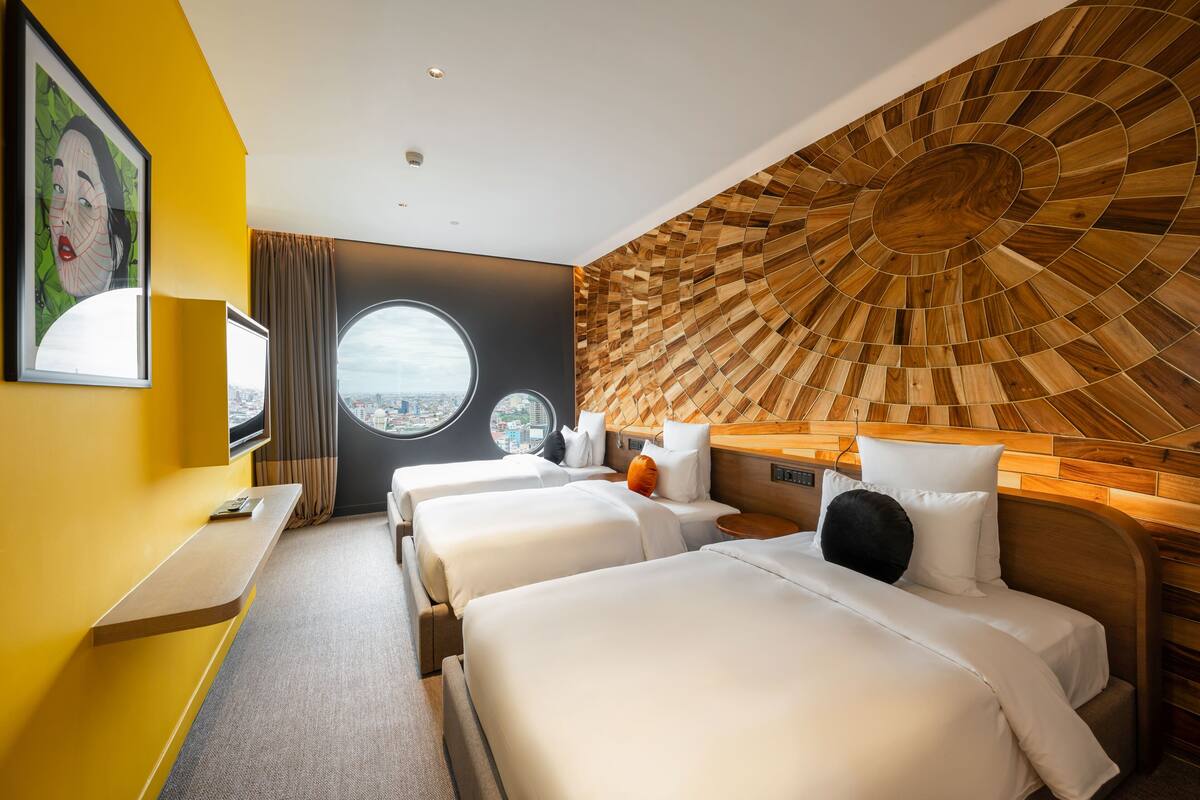 A hotel room with three beds, a city view, a wooden wall design, and a round window.