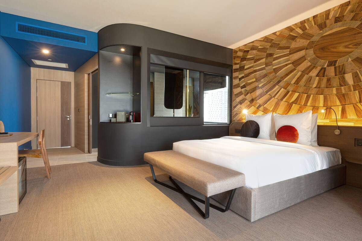 A modern hotel room with a curved wall, a large bed, a bench, and a unique wooden ceiling design.