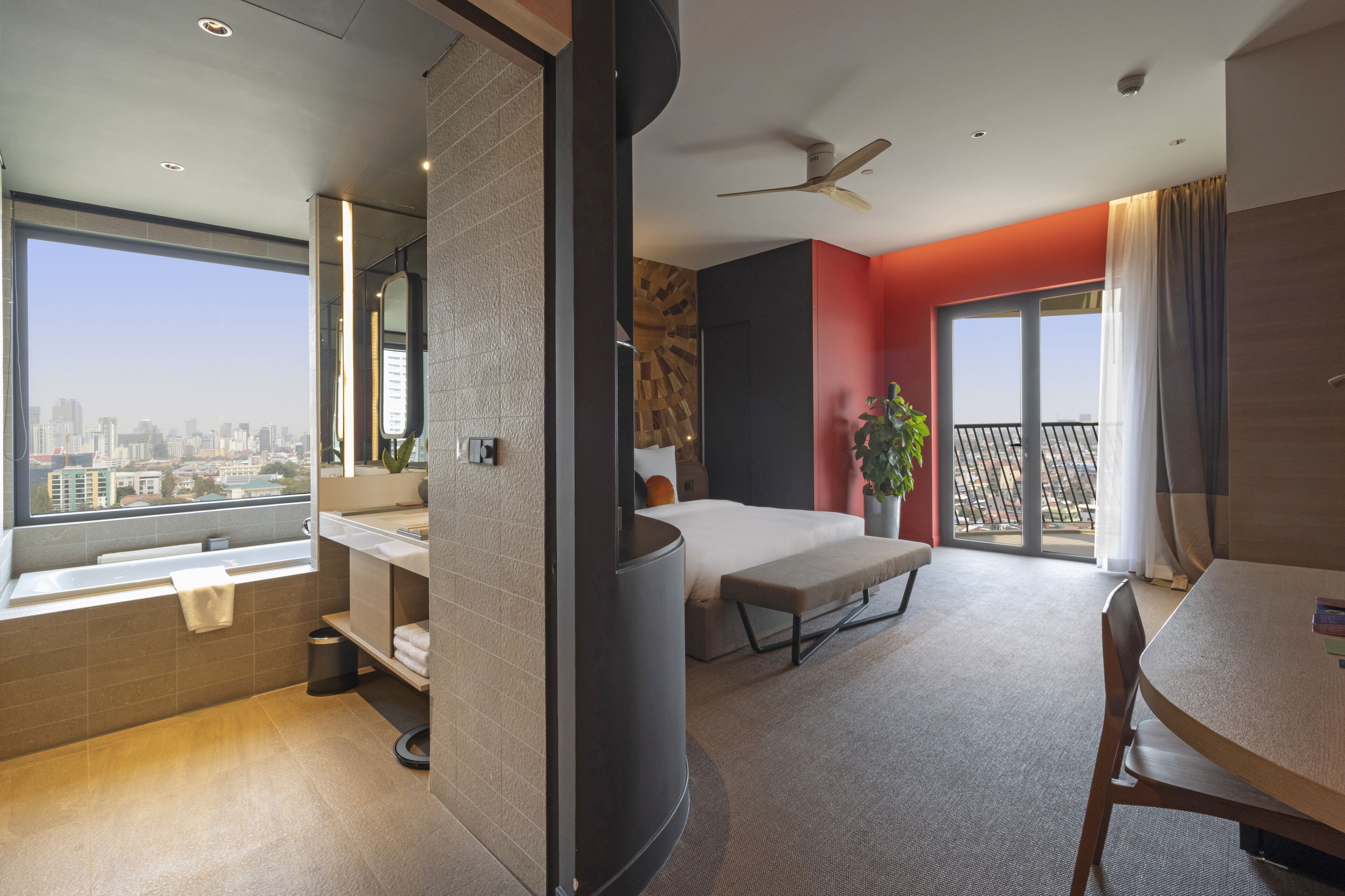 A modern hotel room with a city view, a balcony, a dining area, and a large bed.