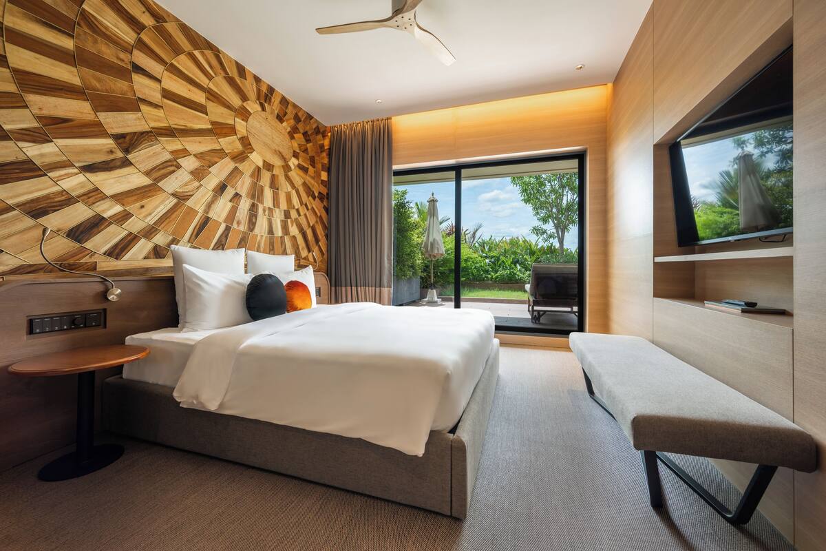 A modern hotel room with a large bed, a bench, a TV, and a view of greenery outside.