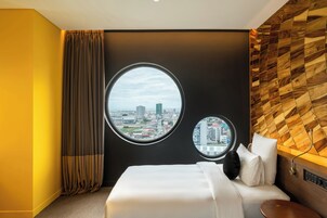 Een hotelkamer met een groot bed, twee ronde ramen met uitzicht op de stad, een houten hoofdbord en een gordijn aan de linkerkant.