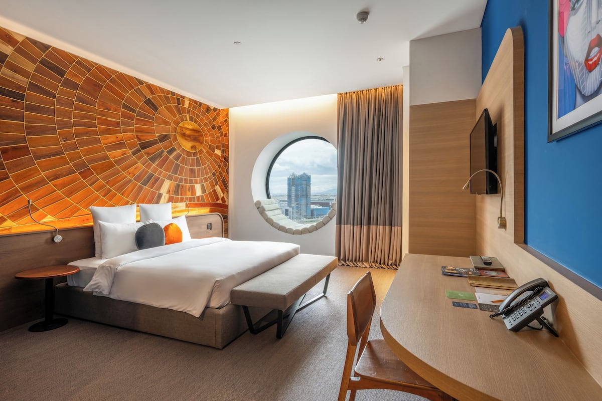 A modern hotel room with a large bed, a round window with a city view, a desk with a phone, and a wall with a geometric pattern.