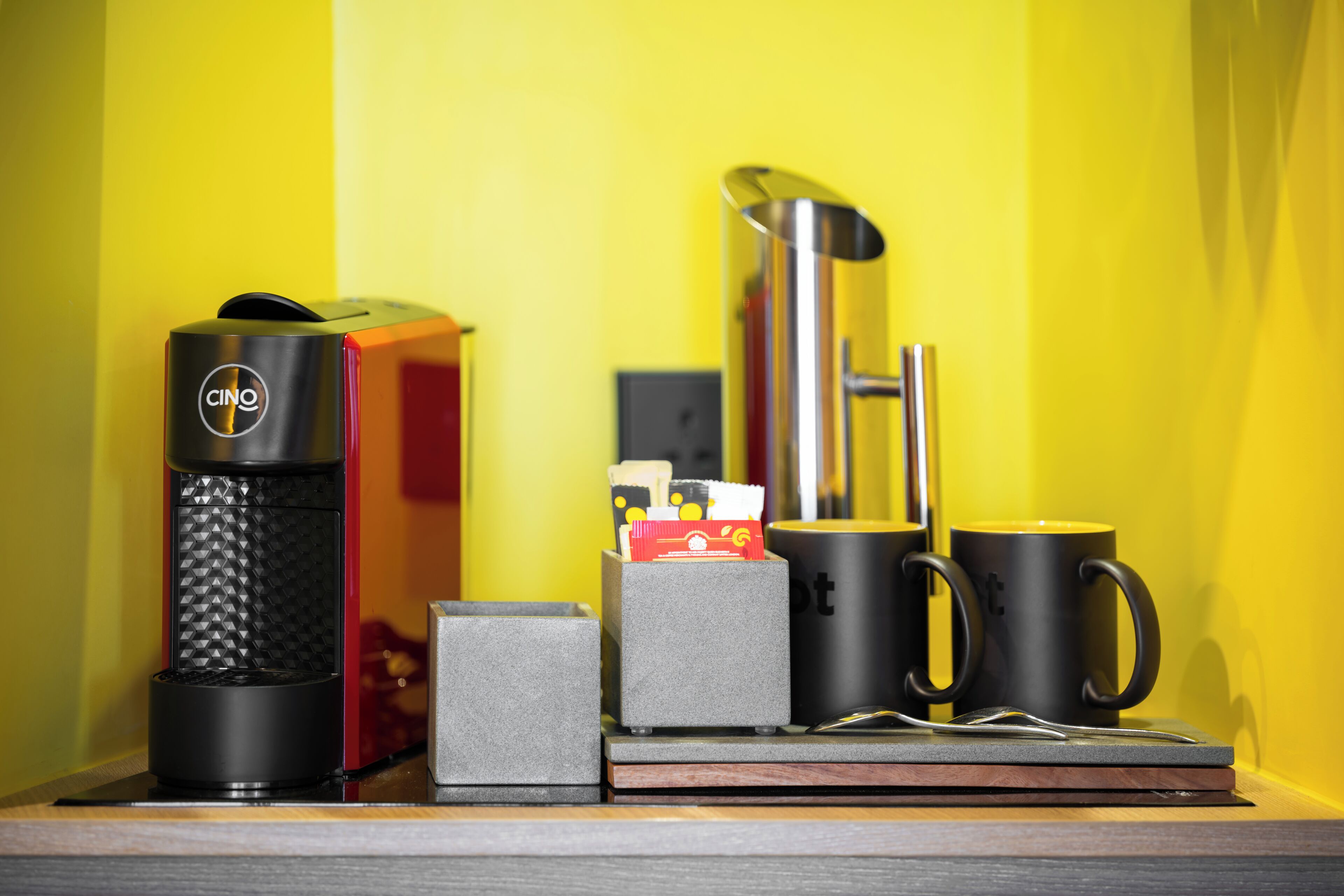 A coffee maker, a box, a mug, and a cup on a wooden surface against a yellow wall.
