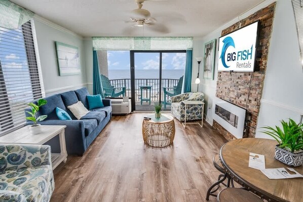 A spacious living room with a large window offering a view of the beach, a blue sofa, a round coffee table, and a dining table with chairs.
