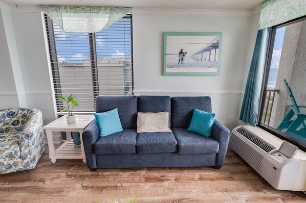 Ein Wohnzimmer mit einem dunkelblauen Sofa, einem gemusterten Sessel, einem kleinen weißen Tisch mit einer Pflanze und einem gerahmten Bild an der Wand.