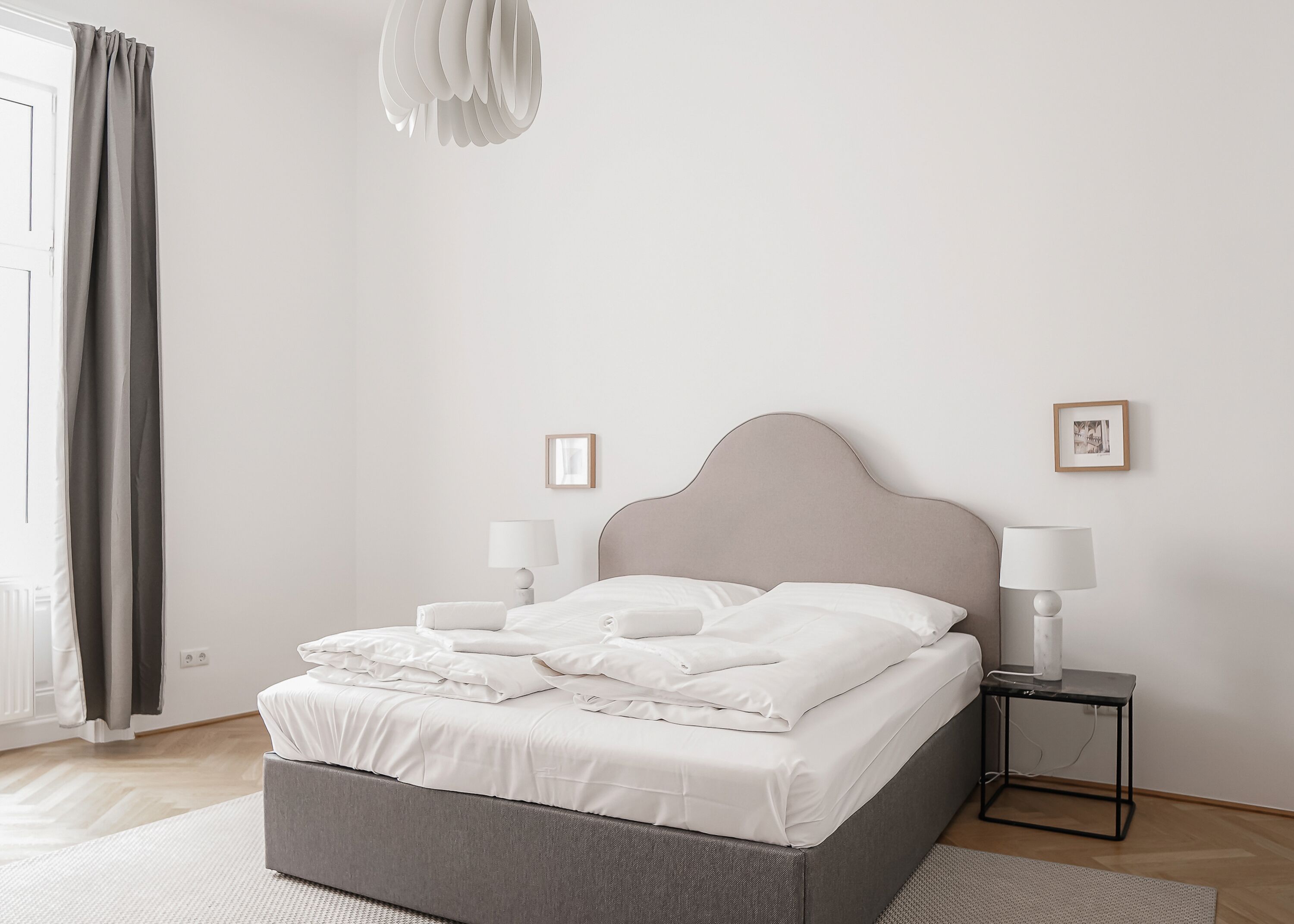 Uma cama bem arrumada com lençóis brancos, duas luminárias de cabeceira e uma mesinha de cabeceira.