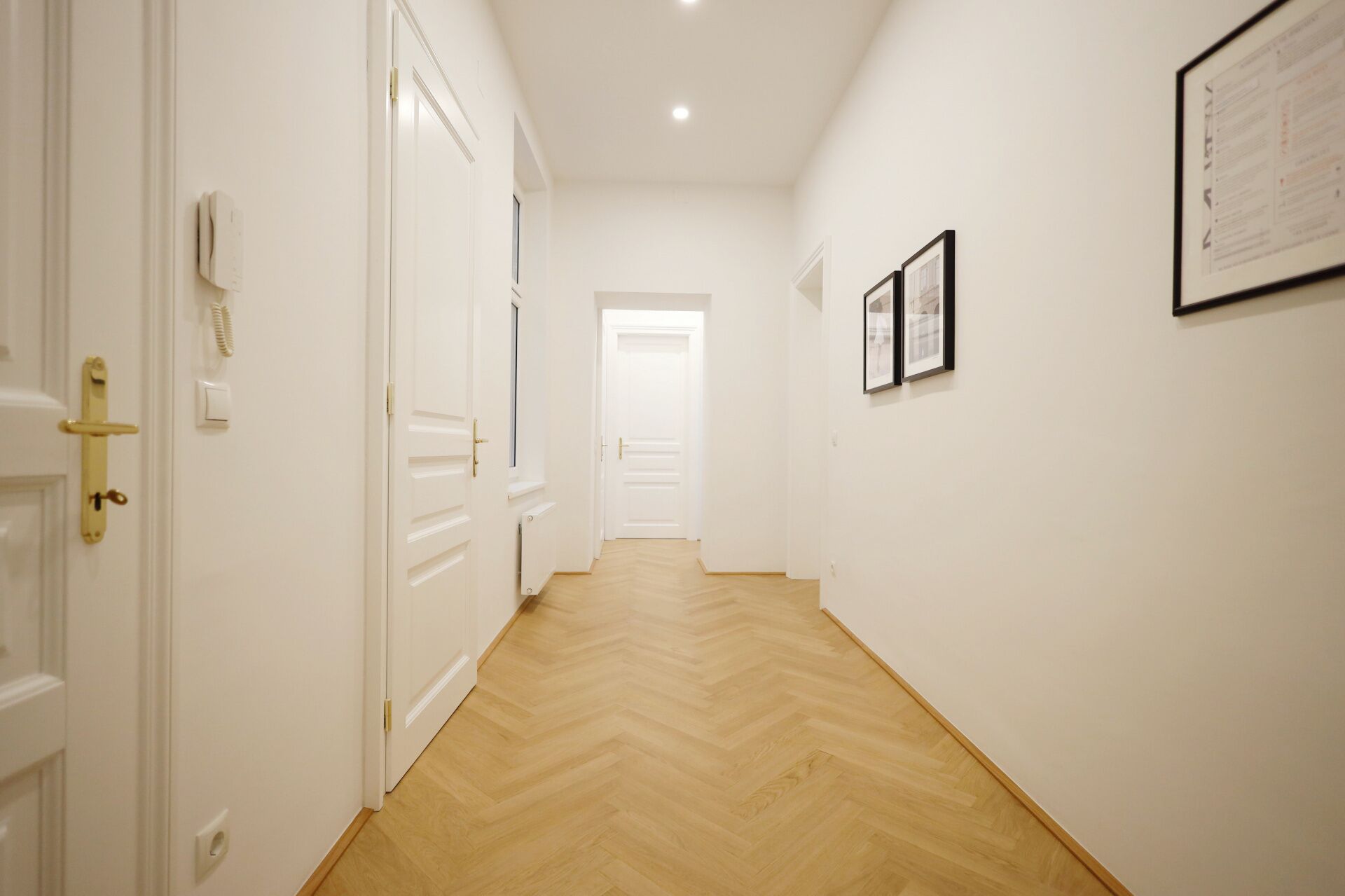 A hallway with white walls and wooden flooring. There are doors on the left, a radiator on the floor, and framed pictures on the wall.