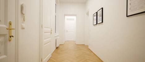 A hallway with white walls and wooden flooring. There are doors on the left, a radiator on the floor, and framed pictures on the wall.