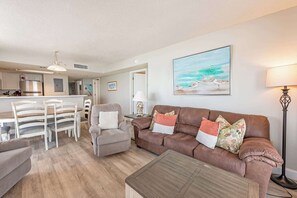 A living room with a brown sofa, a dining table, and chairs. There is a lamp, a painting on the wall, and a kitchen in the background. - Sand Dunes Resort,Remodeled,Pools,HIGH 17th Floor! (Myrtle Beach)