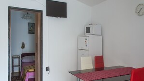Ett litet, väl upplyst rum med ett trägolv, ett vitt kylskåp, en mikrovågsugn, en klocka, ett bord med en röd duk och en röd stol.
