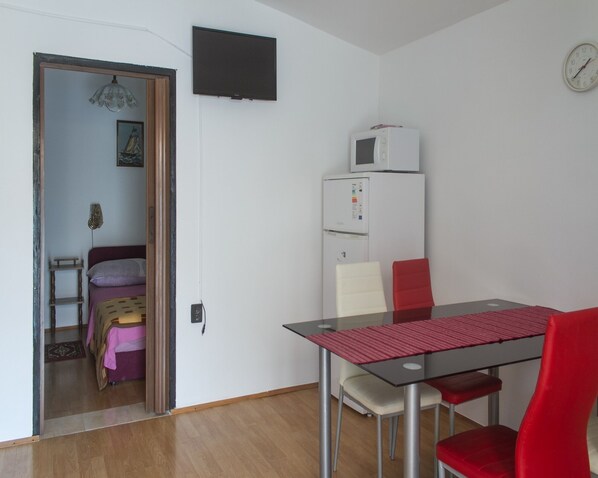 A small, well-lit room with a wooden floor, a white refrigerator, a microwave, a clock, a table with a red tablecloth, and a red chair.