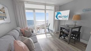 Un salon moderne avec un canapé gris, un téléviseur, une petite table avec une lampe et un balcon offrant une vue agréable.
