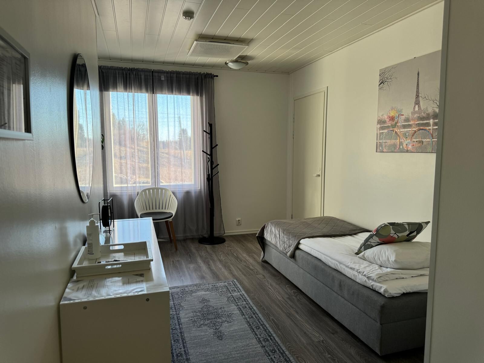 A bedroom with a bed, a desk, a chair, a window with curtains, a coat rack, and a painting of the Eiffel Tower.