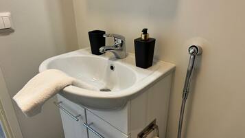 Un baño con lavamanos blanco, grifo, dispensador de jabón, toalla, porta papel higiénico y cabezal de ducha.