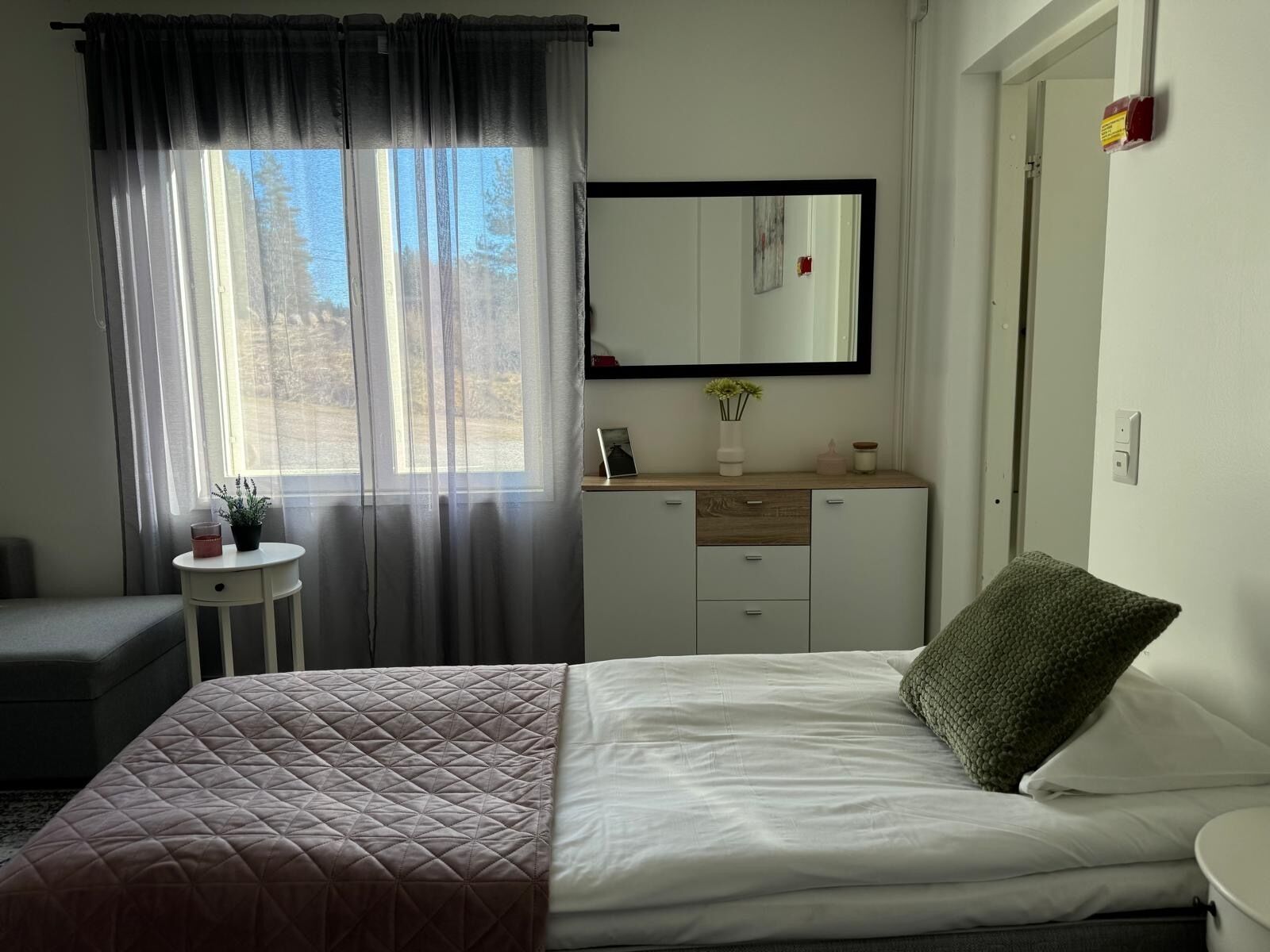 A bedroom with a bed, a bedside table, a dresser, a mirror, and a window with curtains.