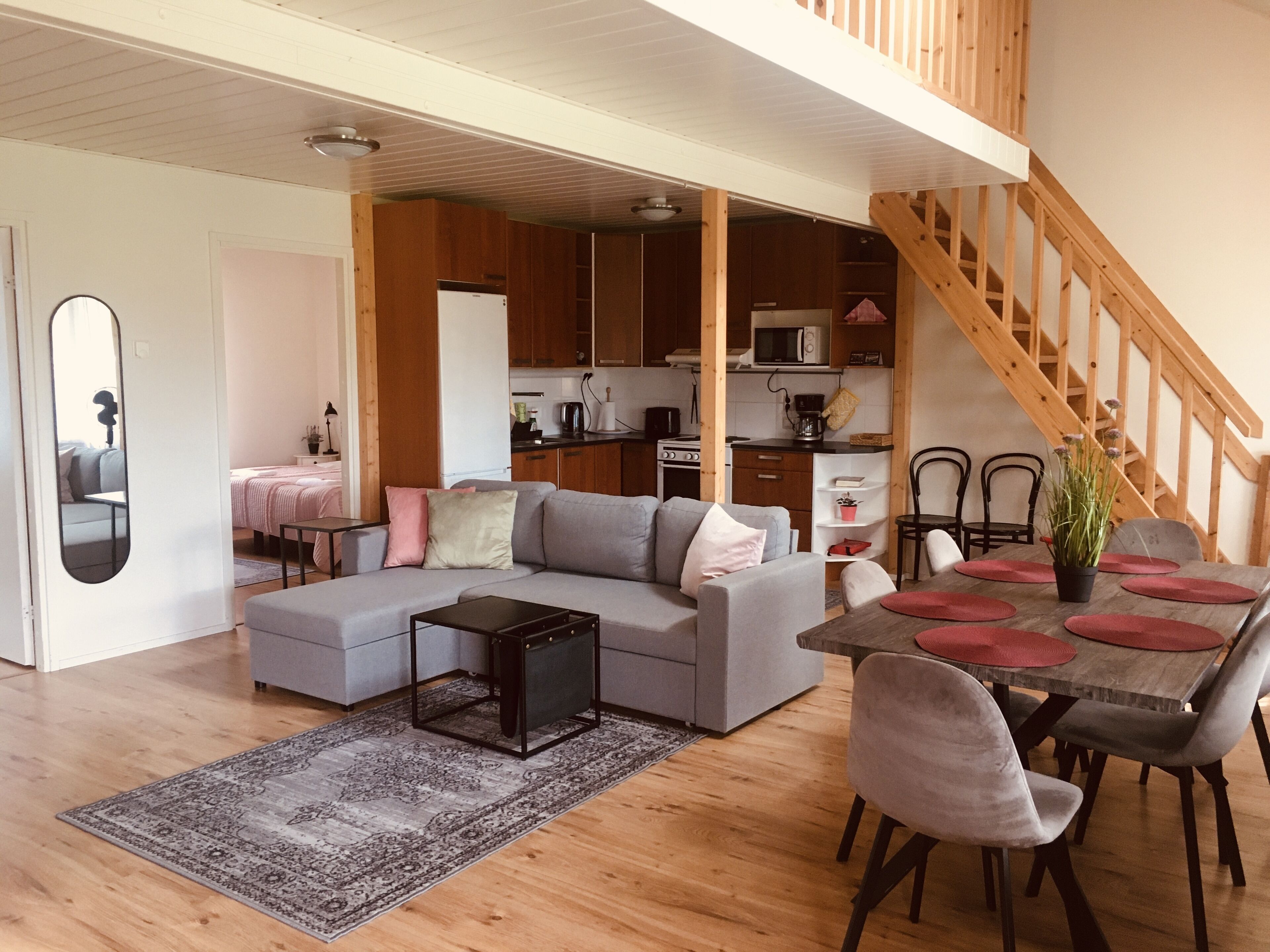 A modern living room with a grey sectional sofa, a small coffee table, and a dining area with a wooden table and chairs. A kitchen with wooden cabinets and a staircase is visible in the background.