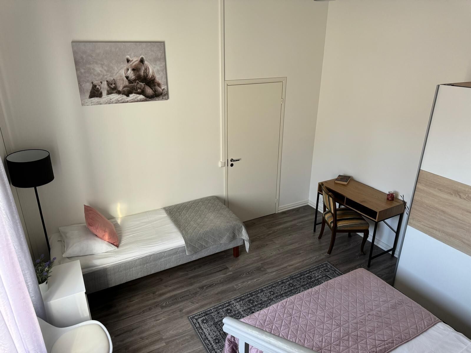 Una habitación pequeña y ordenada con una cama, un escritorio y una silla. Se ve una puerta y un cuadro en la pared.