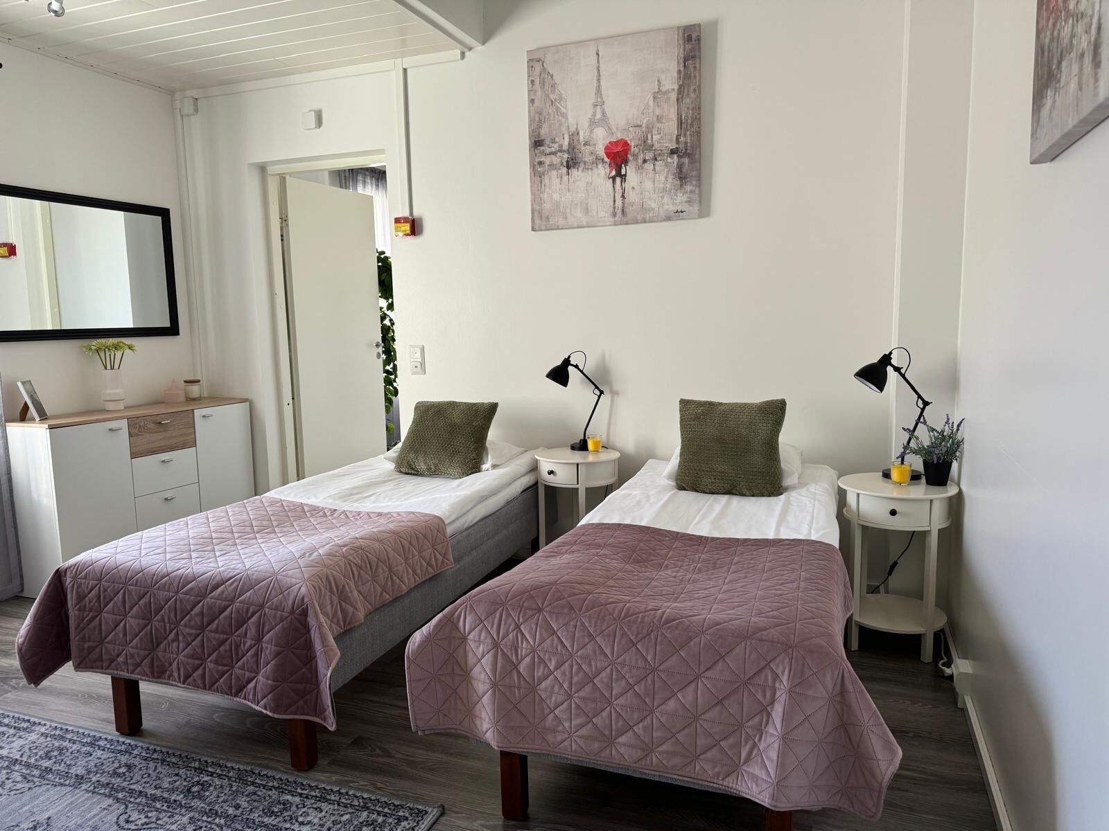 Un dormitorio con dos camas, una mesita de noche con lámpara, un espejo y un cuadro de la Torre Eiffel.
