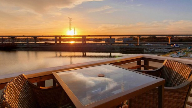 Une terrasse avec une table en verre et des chaises en osier, d’où l’on peut admirer un coucher de soleil sur un pont et l’eau.