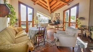 A spacious living area with a high wooden ceiling, a dining table with chairs, a sofa, and a white armchair. There are large windows allowing natural light and a view of the outdoors. - Villa With Large Garden Close to the Sea, Between Etna and Taormina (Acireale)