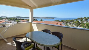 A balcony with a table and chairs, overlooking a coastal view with buildings and trees. - Apartments Jaklin (Rab)