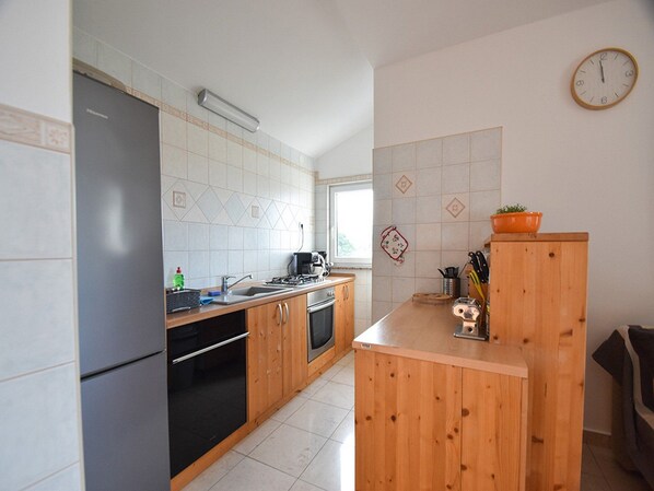Une cuisine équipée d’un réfrigérateur, d’un four, d’un évier et de meubles de rangement en bois. Il y a une horloge au mur et une fenêtre qui laisse entrer la lumière naturelle.