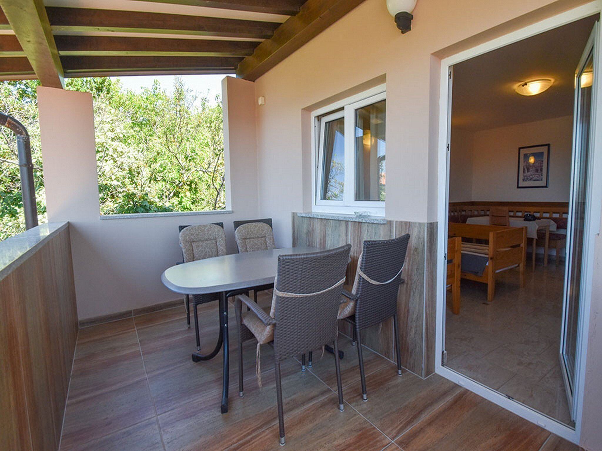 Ein überdachter Patio-Bereich mit Tisch und Stühlen, einem Fenster und Blick ins Grüne.