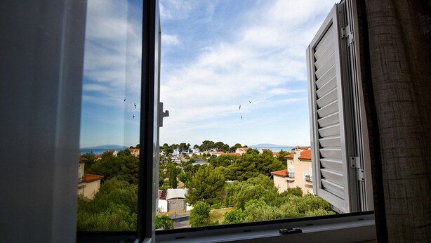A window with a view of a residential area, including houses and trees. - Apartments Jaklin (Rab)