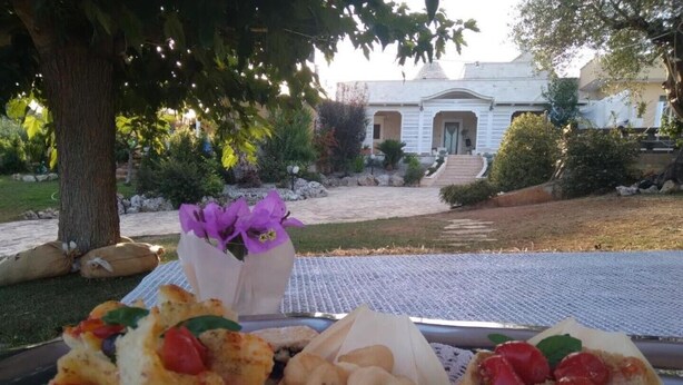 A table with food items and a vase of purple flowers, set outdoors with a building in the background. - Trullo Relais "La Perla Bianca" (Contrada Pozzomasiello-pentimi Locorotondo)