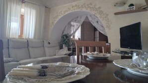 A living room with a sofa, a dining table with plates, a TV, and a decorative archway. - Trullo Relais "La Perla Bianca" (Contrada Pozzomasiello-pentimi Locorotondo)