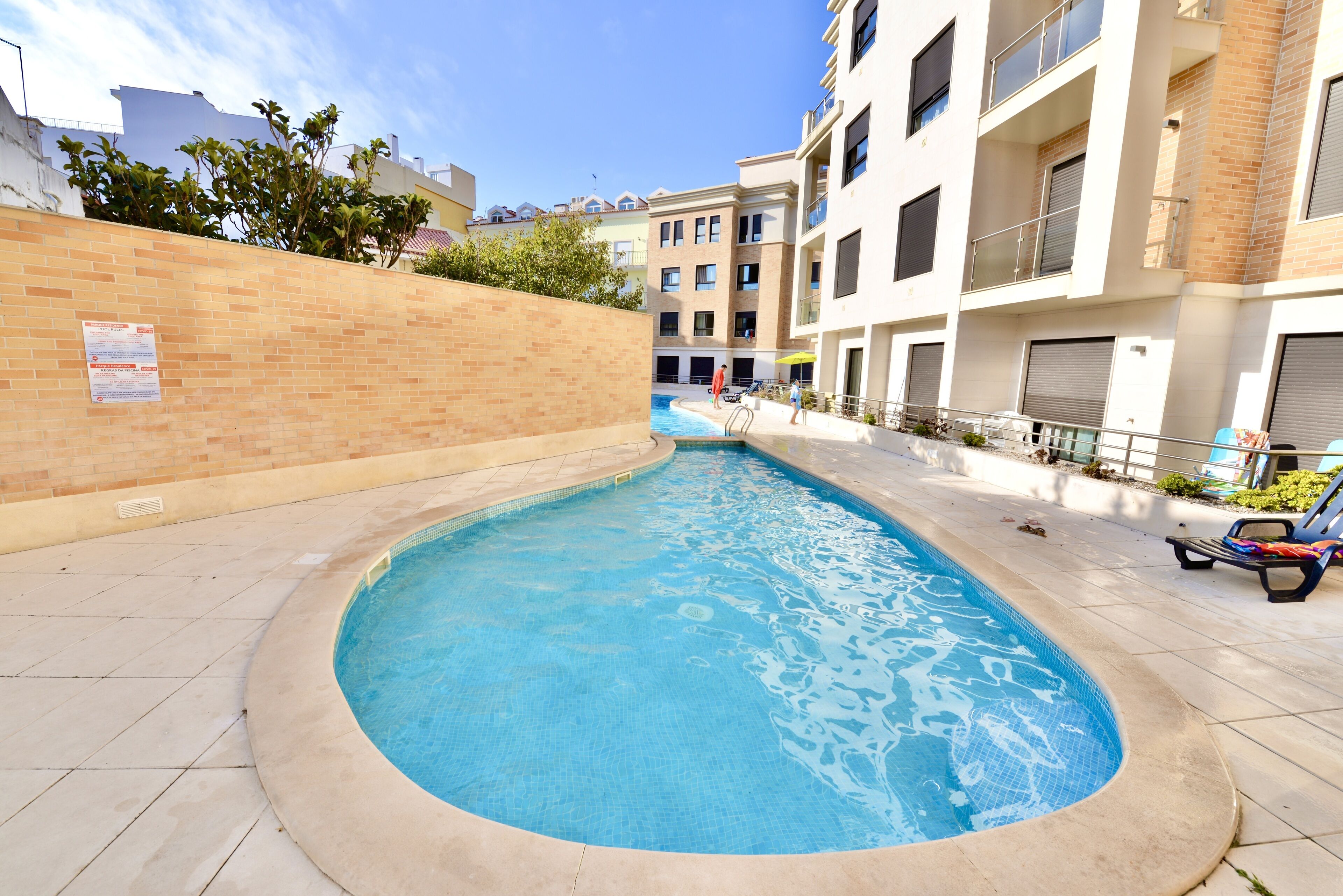 A swimming pool with a tiled edge, surrounded by a brick wall and a building with balconies.
