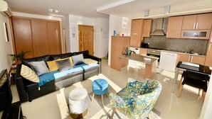 Una sala moderna con un sofá negro, un reposapiés azul y un sillón colorido. Se ve una cocina con muebles de madera y electrodomésticos de acero inoxidable al fondo.