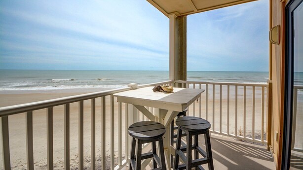 Un balcon avec une table et deux tabourets donnant sur une plage.