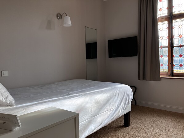 A bedroom with a single bed, a bedside table, a wall-mounted TV, a mirror, and a window with curtains. - Le Château d'Aubry (Aubry-du-Hainaut)