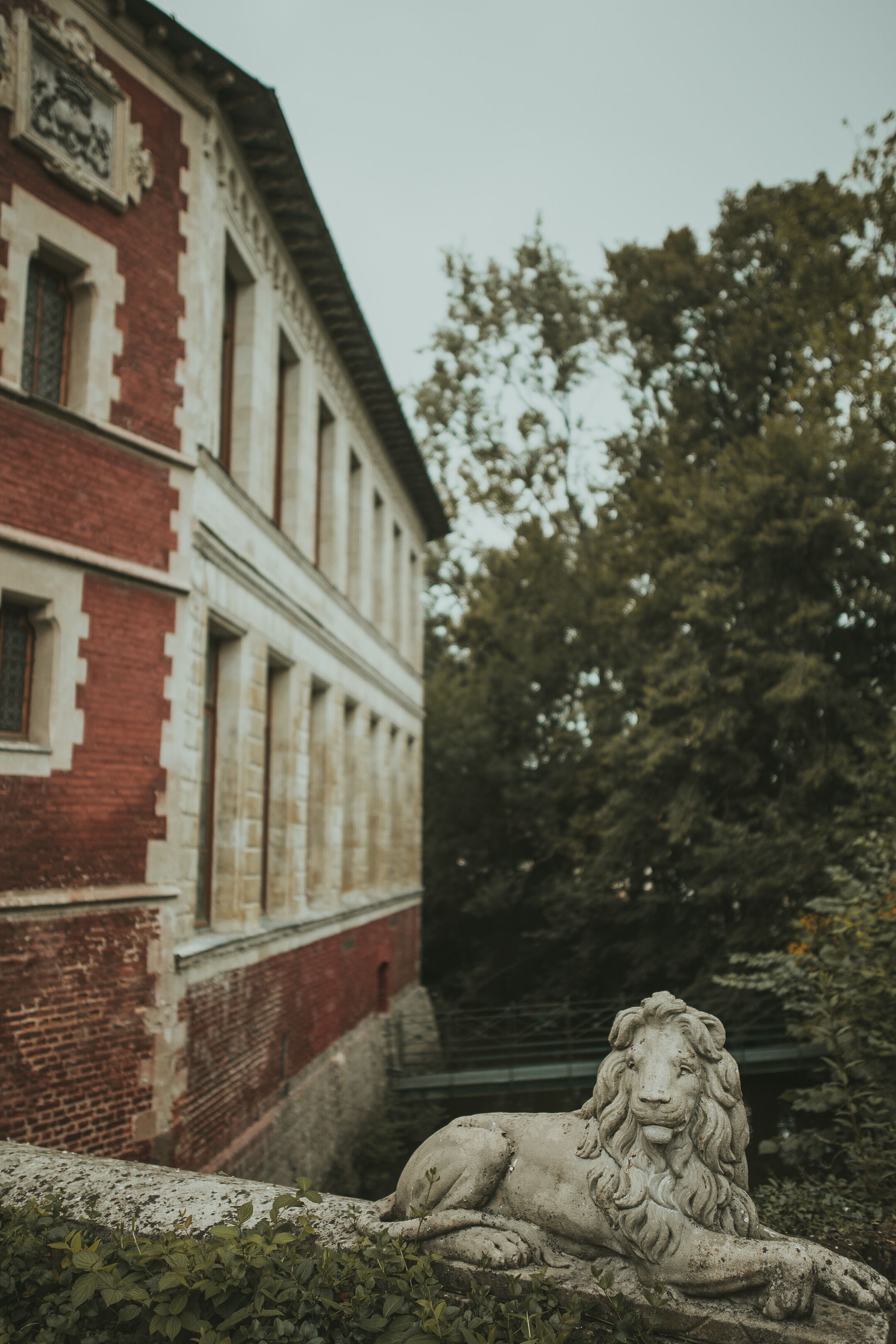 Una estatua de león de piedra frente a un edificio de ladrillo rojo con ventanas grandes y una escalera que conduce hacia él.