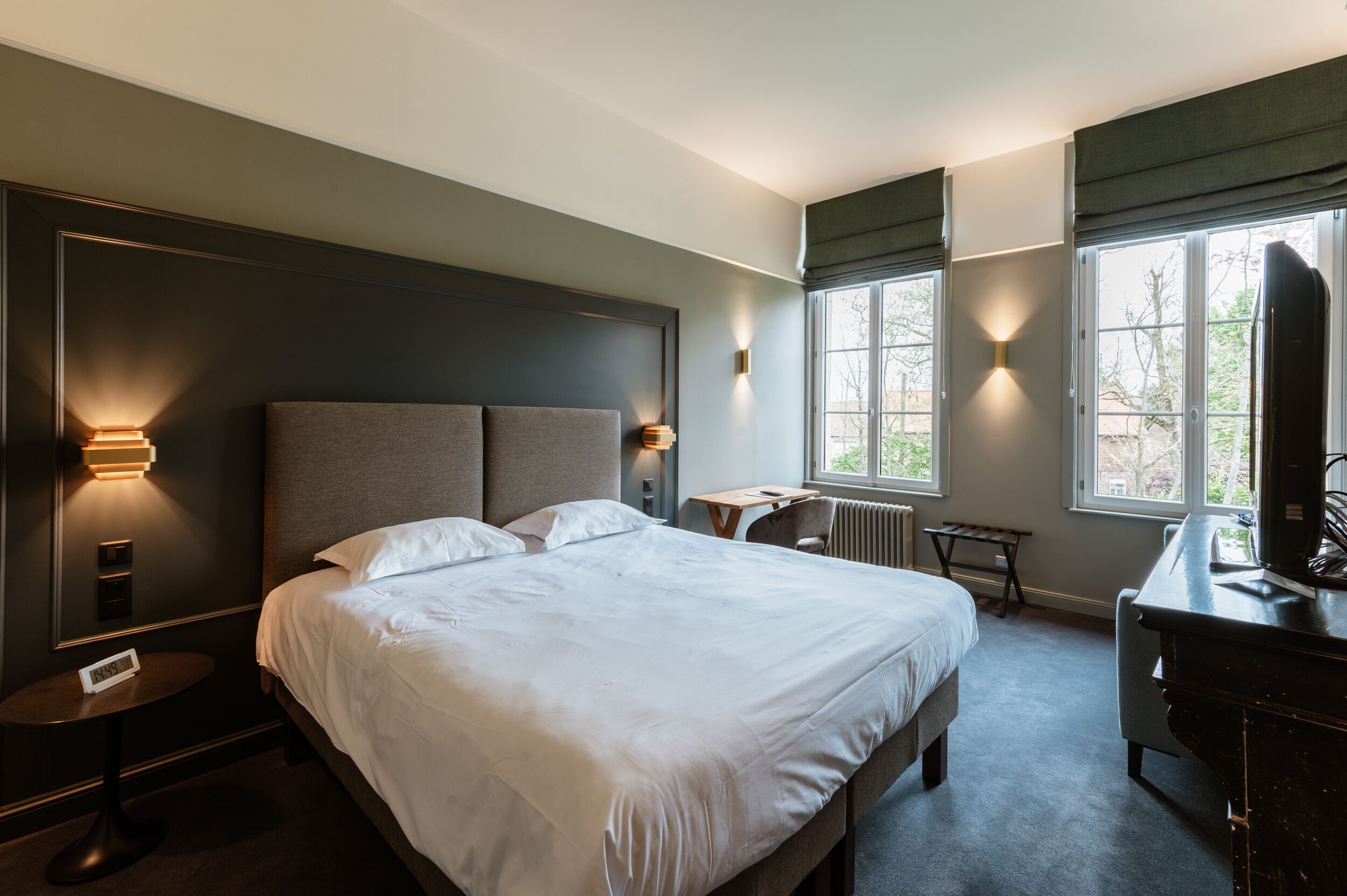 A modern hotel room with a large bed, bedside tables, a desk, and a view of the outdoors through large windows.