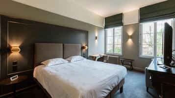 A modern hotel room with a large bed, bedside tables, a desk, and a view of the outdoors through large windows.
