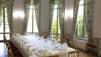 A dining room with a long table set for a meal, flanked by tall windows with curtains, wooden chairs, and a chandelier.
