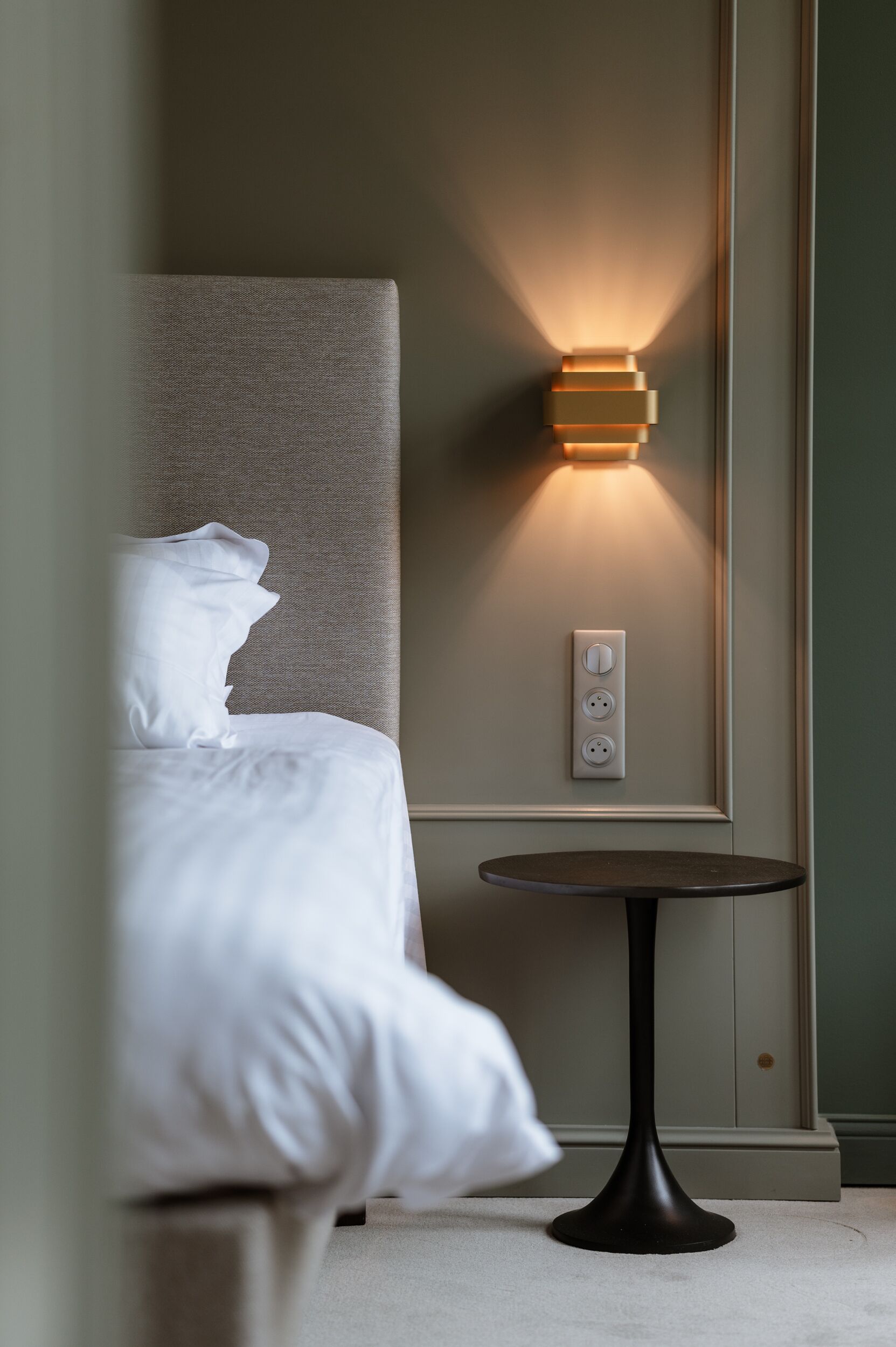 A neatly made bed with a headboard, a wall-mounted light fixture, and a small round table.