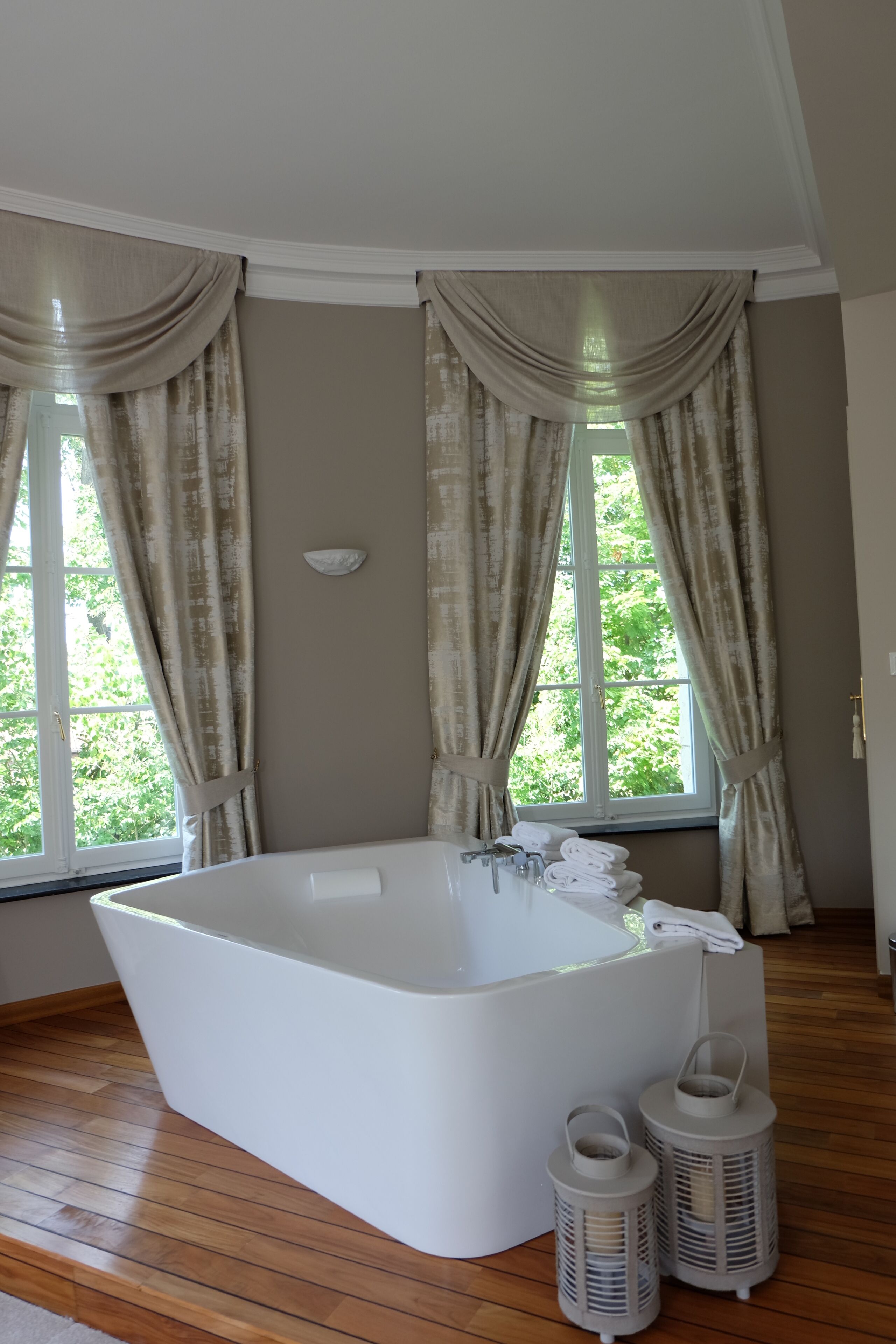 Una grande vasca da bagno bianca, indipendente, in una stanza con pavimento in legno, ampie finestre con tende color beige e due lanterne bianche.