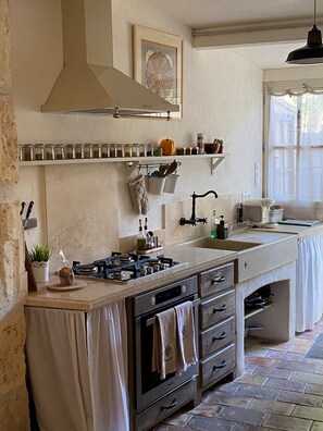 A kitchen with a gas stove, oven, and a sink. There are cabinets, a hanging pot rack, and a window with curtains.