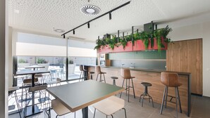 A modern restaurant with a high ceiling, large windows, and a bar area with stools. -  Livensa Living Studios Madrid Alcobendas (Alcobendas)